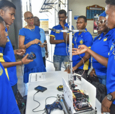 Students gathered around a robot. Photograph published by NY Carib News and Caribbean National Weekly. No photo credit was printed.